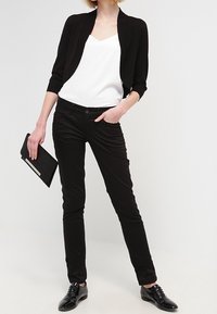 Černý krátký kardigan, bílý top s výstřihem do V, černé přiléhavé kalhoty a černé šněrovací boty. Modelka drží černé psaníčko se stříbrným doplňkem.