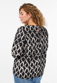 Blouse noire avec un motif de feuilles texturé beige, dotée d'un col rond et d'un dos en trou de serrure avec une fermeture par lien.