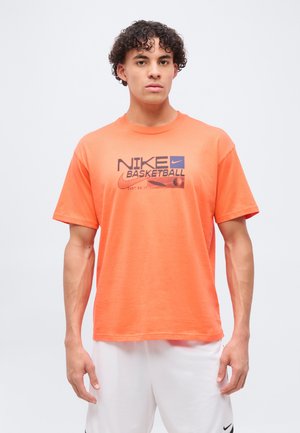 Jeune homme aux cheveux bouclés portant un t-shirt de basketball Nike orange et un short Nike blanc, debout devant un fond clair uni.