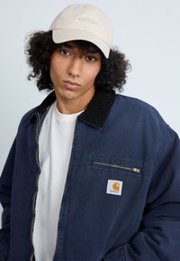 Mornarska zip majica s ovratnikom od kordura, sa prednjim džepom i Carhartt logom. Nosena preko bijele majice i uparena s bež kapom.