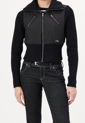 Person trägt schwarzen, gerippten Reißverschluss-Pullover mit gepolstertem Oberkörperbereich und Reißverschlusstaschen, kombiniert mit dunkler Jeans.