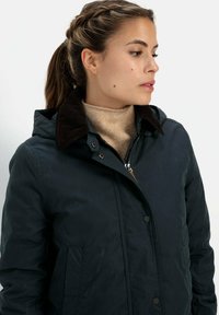 Donkergroene waterdichte jas met een corduroy kraag, drukknoopsluitingen, zijzakken en een capuchon. Draag je over een beige gebreide trui.