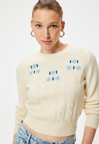 Krämbelagd kabelstickad tröja med blå blombroderier och gröna inslag på bröstet. Ribbad nederkant och ärmslut ger textur och detaljer.