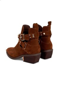 Stivali alla caviglia in suede marrone con punta rotonda, tacco spesso e fibbie decorative. Tra le caratteristiche ci sono borchie e un accento in legno a contrasto sul tacco.