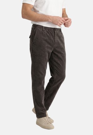 Uomo con pantaloni in velluto a coste grigio scuro, t-shirt bianca e sneakers beige, in piedi con le mani vicino alla vita su sfondo bianco.