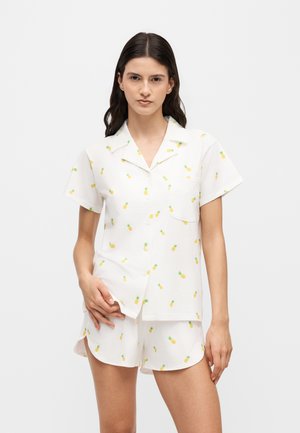Jeune femme portant une chemise blanche à manches courtes boutonnée et un short assorti avec de petits imprimés ananas jaunes sur un fond uni.