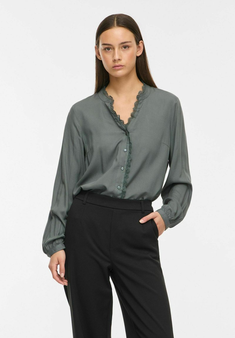 Femme aux longs cheveux foncés porte un chemisier vert foncé boutonné avec bordure en dentelle et un pantalon noir taille haute, posant avec une main dans la poche.
