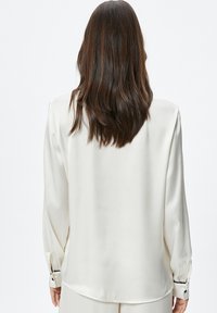 Blusa de satén blanca con mangas largas, corte holgado y detalles de botones negros en los puños. Textura suave y dobladillo redondeado en la parte posterior.