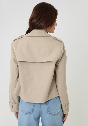 Frau mit langen braunen Haaren, die einen beigen, kurz geschnittenen Trenchcoat und hellblaue, hoch taillierte Jeans trägt, mit dem Rücken zur Kamera gewandt.