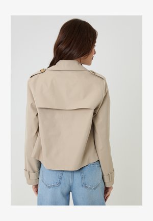 Frau mit langen braunen Haaren, die einen beigen, kurz geschnittenen Trenchcoat und hellblaue, hoch taillierte Jeans trägt, mit dem Rücken zur Kamera gewandt.