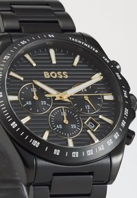Montre chronographe en acier inoxydable noir avec un cadran sombre strié, des index horaires dorés, trois sous-cadrans et une échelle tachymétrique sur la lunette.