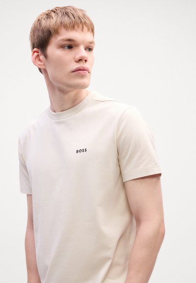 T-shirt beige clair en coton à manches courtes, avec un col rond et le logo noir "BOSS" sur la poitrine gauche. Texture lisse.