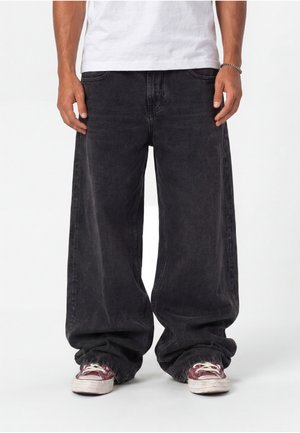 Homme portant un jean noir oversize et des baskets rouges avec des lacets blancs, debout devant un fond blanc uni.