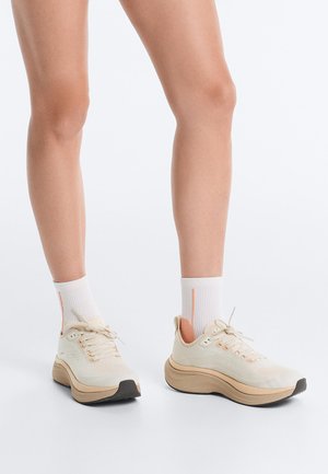 Noge v belih in bež masivnih supergah z belimi nogavičkami s oranžnimi črtami, ki stojijo na enobarvni beli površini.