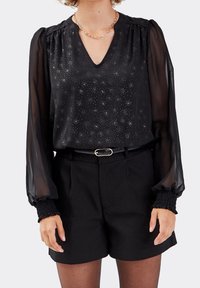 Blouse noire à manches longues transparentes, décolleté en V et paillettes métalliques. Associée à un short noir taille haute et une ceinture noire.