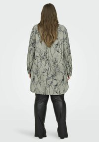 Tunique verte longue avec imprimé botanique noir, associée à un pantalon en cuir synthétique noir. La tunique a une coupe décontractée et des manches longues.