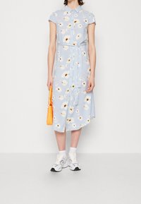 Ljusblå, kortärmad skjortklänning med blommönster och knappar, knut vid midjan, parat med vita strumpor och grå sneakers, håller en orange väska.