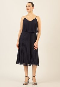 Marineblaues, plissiertes Kleid mit V-Ausschnitt und verstellbaren Spaghettiträgern. Mit einem geschichteten Oberteil, fließendem Rock und schwarzen, mit Schleifen verzierten, hohen Sandalen.