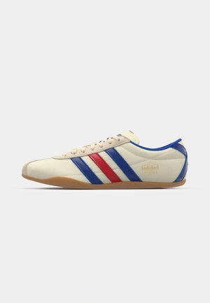 La sneaker presenta una toma sintetica crema con strisce blu e rosse, una suola in gomma beige e un collare imbottito. Logo "Adidas Tokyo" sul lato.