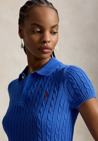 Femme aux cheveux tressés portant des boucles d'oreilles en coquillage et un polo en maille torsadée bleu, regardant vers le bas et vers sa gauche.