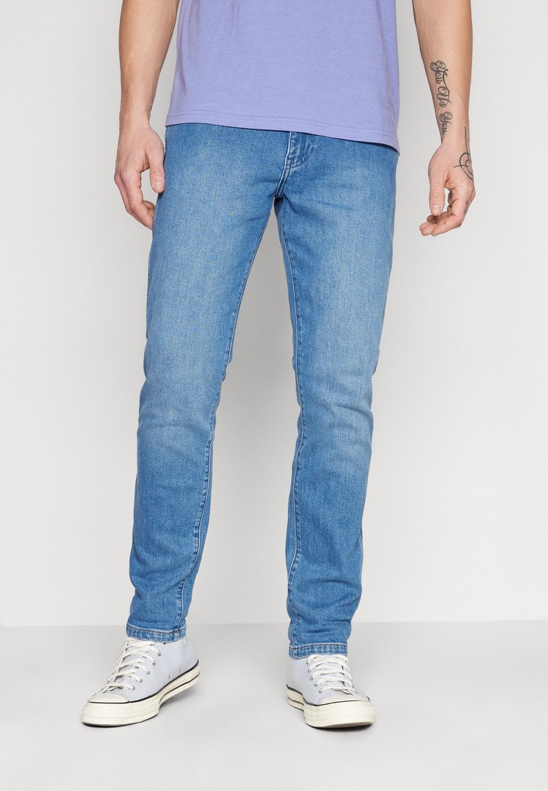 Man som bär ljusblå slim-fit-jeans och vita sneakers, står mot en neutral ljus bakgrund, med händerna avslappnat vid sidorna.