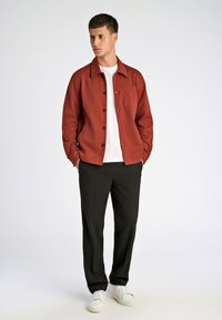 Rotes Hemd mit Knopfleiste, einer linken Brusttasche, schwarzen Knöpfen und lockerer Passform, kombiniert mit schwarzen Hosen und weißen Sneakern.
