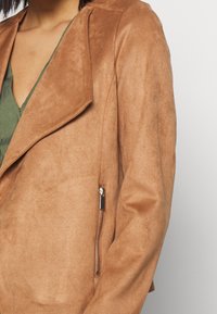 Donna che indossa una giacca di suede marrone con una tasca con zip, sopra un top verde a scollatura a V, mostrata dalle spalle fino a metà torso.