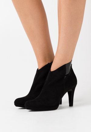 Schwarze Wildleder-Stiefeletten mit spitz zulaufender Zehe, hohem Absatz und ausgeschnittenem Design mit glänzenden schwarzen Akzenten an den Seiten.