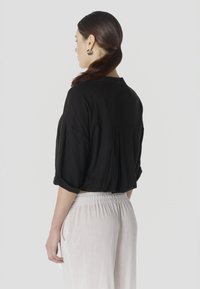 Žena s tmavými vlasmi zviazanými v nízkych copoch, oblečená v čiernej voľnej blúzke s vyhrnutými rukávmi a svetlých nohaviciach s vysokým pásom.