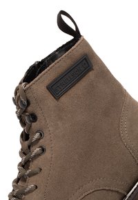 Brązowe zamszowe ankle boot z czarną metką logo, wyposażone w okrągłe sznurowadła i metalowe oczka. Projekt zawiera akcenty w postaci przeszyć oraz pętelkę do zakładania.