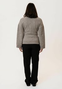 Šedý pletený svetr s širokými rukávy a elastickým lemem, nošený s černými kalhotami. Materiál vypadá měkce a texturovaně. Jednoduché pozadí.