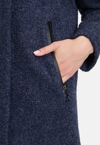 Granatowy płaszcz wełniany z teksturowanym wykończeniem, z boczną kieszenią na zamek i czarnym sprzętem zamkowym. Ręka oparta w kieszeni.