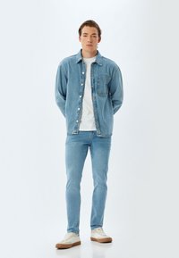 Chaqueta de mezclilla azul claro con un bolsillo frontal, camiseta blanca debajo, jeans azul claro a juego y zapatillas beige con suelas de goma.