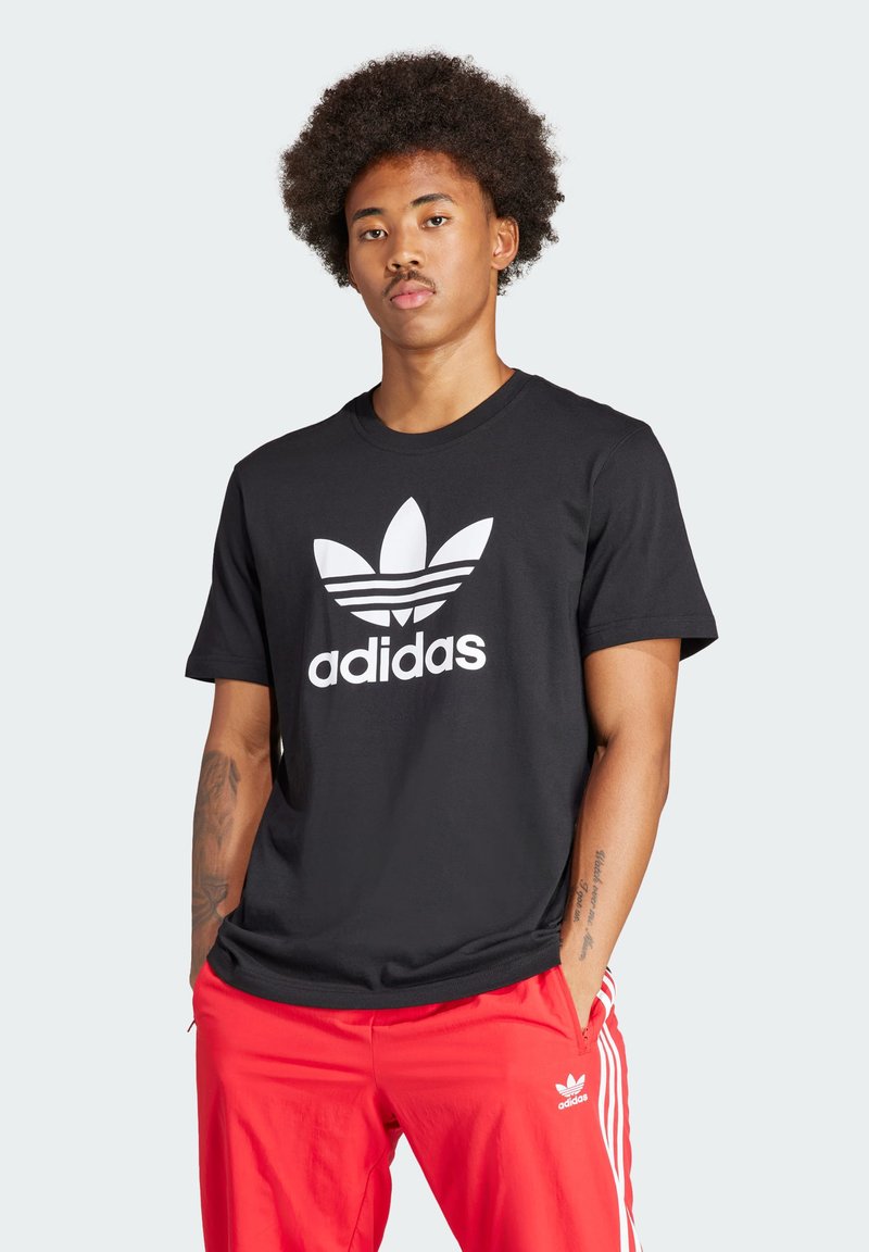 Zwarte katoenen t-shirt met een groot wit Adidas-logo en drie strepen op de voorkant, gecombineerd met rode trainingsbroek met witte accenten.