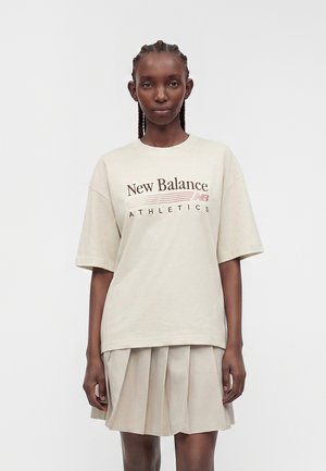 Γυναίκα με πλεξούδες, που φοράει μια μπεζ oversize μπλούζα New Balance Athletics και μια μπεζ πλισέ φούστα, στέκεται μπροστά σε ένα απλό φόντο.