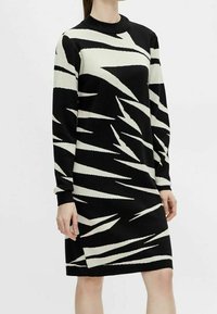 Robe en maille rayée noire et crème, dotée d'un col rond, de manches longues et d'une coupe droite avec une fente latérale pour faciliter les mouvements.