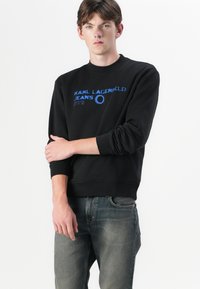 Sudadera negra con texto azul, cuello redondo, puños y dobladillo acanalados. Se lleva con jeans gris desgastados. Presenta una marca en letras en negrita.