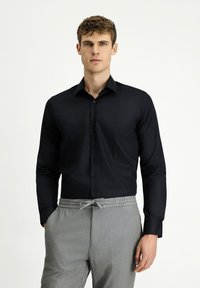 Man wearing a fitted black button-up shirt and light gray drawstring pants, standing with hands in pockets against white background.