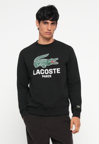 Juodas medvilninis džemperis su žaliu krokodilo grafiniu atvaizdu ir baltais užrašais "LACOSTE PARIS". Su gipiūriniais rankogaliais ir apatine dalimi.