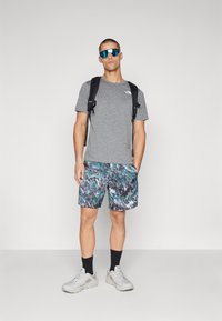 Camiseta gris de manga corta, pantalones cortos a rayas en azul y verde, mochila negra, gafas de sol reflectantes azules, zapatillas grises y calcetines negros.