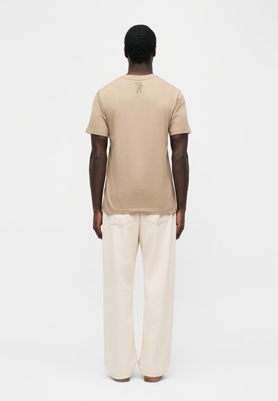 Άνδρας με μπεζ κοντομάνικο μπλουζάκι με μικρό γραφικό αστροναύτη στο πάνω μέρος της πλάτης και χαλαρό παντελόνι σε εκρού χρώμα, γυρισμένος προς τα πίσω σε λευκό φόντο.