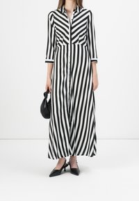 Robe maxi à rayures noir et blanc avec des manches longues, un boutonnage à l'avant et une silhouette cintrée. Portée avec un sac noir et des escarpins à bout pointu.