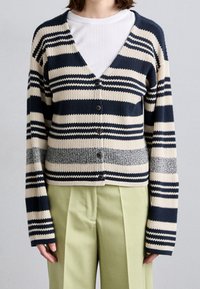 Striped cardigan in navy and cream with ribbed texture, button closure, and long sleeves. Worn over a white shirt and light green trousers.
