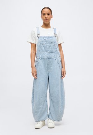 Femeie în picioare, purtând salopetă largă cu dungi subțiri albastre deschis peste un tricou alb cu mâneci scurte și pantofi sport albi, pe un fundal simplu.