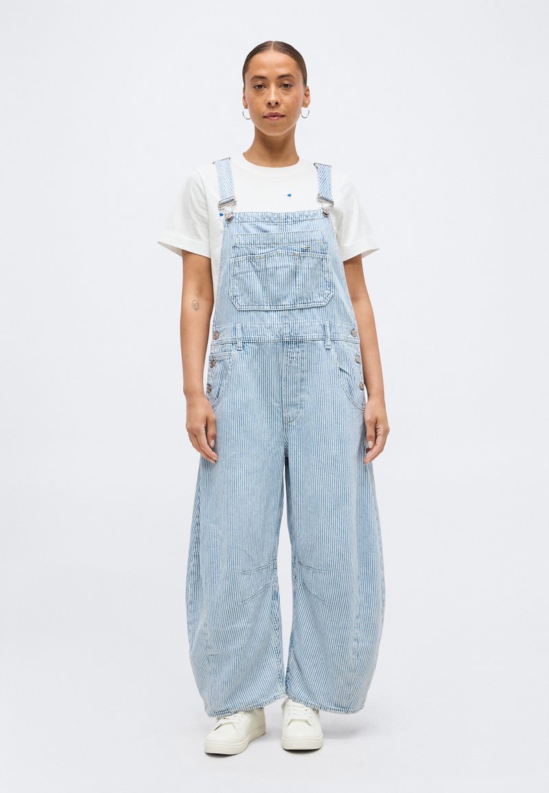 Γυναίκα όρθια, φορώντας ανοιχτό μπλε ριγέ ολόσωμη φόρμα με φαρδιά μπατζάκια πάνω από λευκό κοντομάνικο πουκάμισο και λευκά αθλητικά παπούτσια, μπροστά σε απλό φόντο.