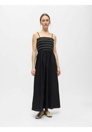 Femme debout portant une robe maxi noire sans manches avec un corsage smocké et des ballerines beiges sur un fond blanc uni.