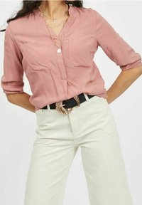 Donna con una blusa rosa a maniche lunghe arrotolate, pantaloni beige a vita alta, una cintura nera con fibbia circolare dorata e collane dorate a strati.