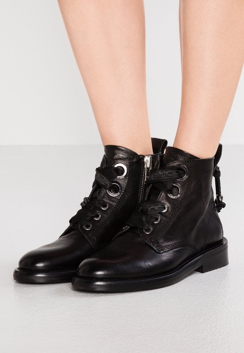 Botas de tobillo de cuero negro con gruesos cordones negros, ojales plateados, cremalleras laterales, usadas sobre piernas desnudas contra un fondo liso claro.