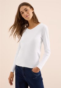 Weißes Langarmhemd mit V-Ausschnitt, aus weichem Stoff. Kombiniert mit dunkelblauen Jeans mit Fronttaschen und Nähtelementen.