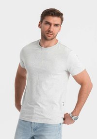 Camiseta corta de manga blanca con un sutil patrón de hojas, escote redondo y una etiqueta en el dobladillo. Combinada con unos vaqueros de color azul claro.